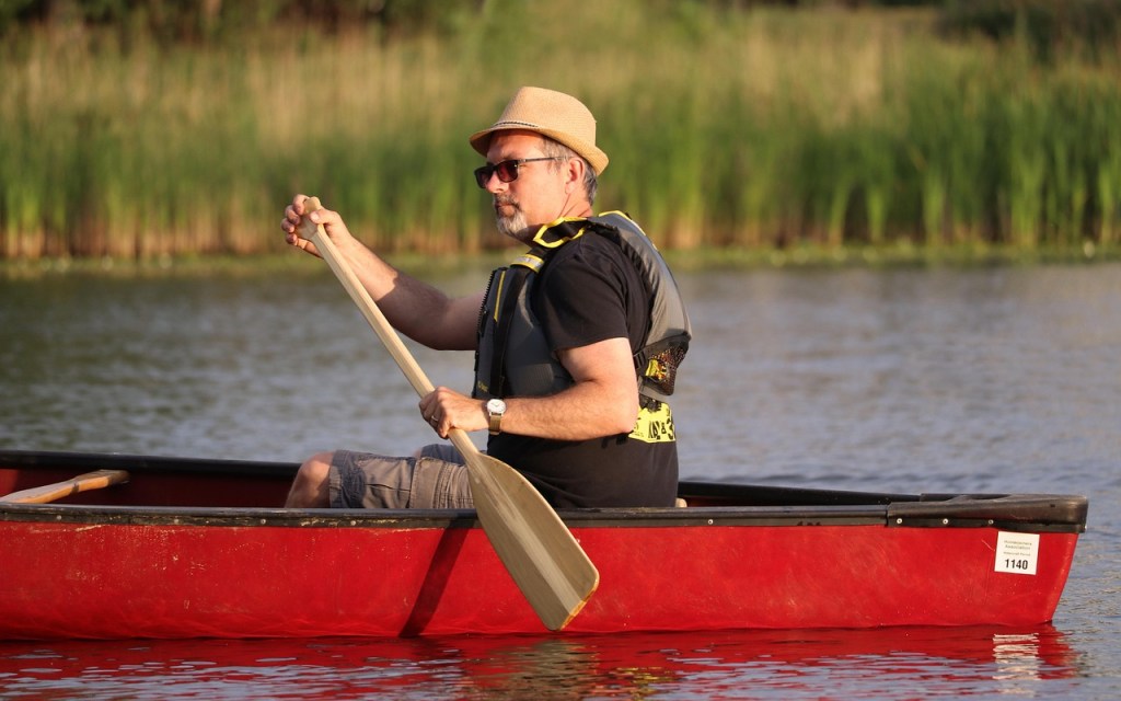 Person paddling a red canoe with a single-bladed oar on a calm lake; image illustrates the difference between canoe oars and double-bladed kayak paddles.
